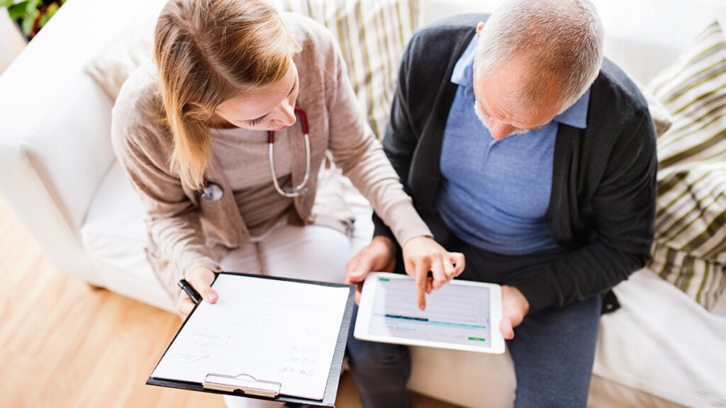 A clinician with a traditional clipboard and paper helps a patient fill out details on a digital tablet.